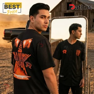 Fuerza Regida x San Francisco Giants This Is Our Dream 2026 Baseball Jersey 1 Man wearing a black Fuerza Regida baseball jersey with orange graphic text and logos, standing outdoors by a mirror.!