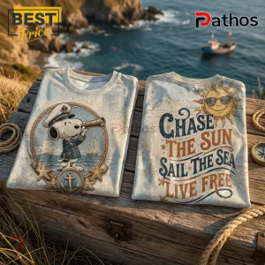 Two folded cotton t-shirts featuring nautical Snoopy graphics, anchor motifs, and ocean-themed typography on wooden crates.!