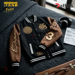 Black and brown wool varsity jacket with gold embroidery, number 3 patch, and striped ribbed trim on dark bedding.!