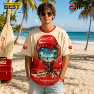 Man wearing a cream and red Coca-Cola t-shirt with a surreal beach-themed graphic and text on a sandy tropical beach.!