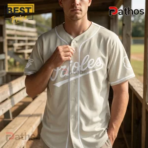 Baltimore Orioles x Ella Langley Limited Edition Baseball Jersey 4 Tan button-up baseball jersey with white Orioles script text and white trim worn by a man at a baseball field dugout.!