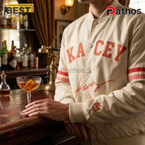 Man wearing cream varsity jacket with orange KACEY lettering, stripes on sleeves, and red signature, inside a vintage bar.!