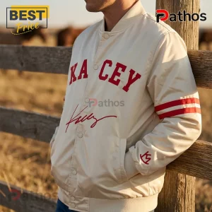 Cream satin varsity jacket with red KACEY lettering, signature embroidery, and striped sleeves near a wooden fence.!